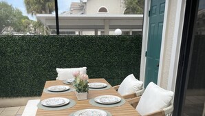 A wooden table set with plates and a vase of flowers, surrounded by white cushioned chairs, on a patio with a green hedge and a building in the background.