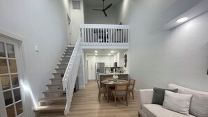 A spacious living area with a staircase leading to an upper level, a dining area with a round table, and a sofa with cushions.