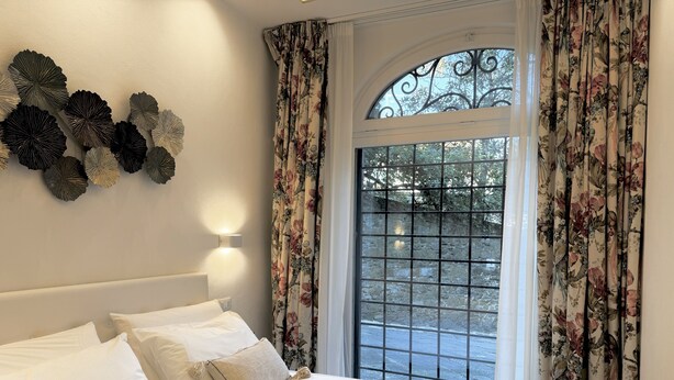 A neatly made bed with white linens and decorative pillows, a wall-mounted light fixture, and a window with floral curtains and a metal grill. - Ponte Vecchio｜Tower of Gelfi (Firenze)