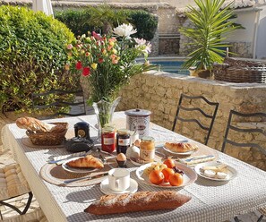 Une table dressée avec du pain, des pâtisseries, des fruits et des condiments, avec en arrière-plan une piscine et de la verdure.