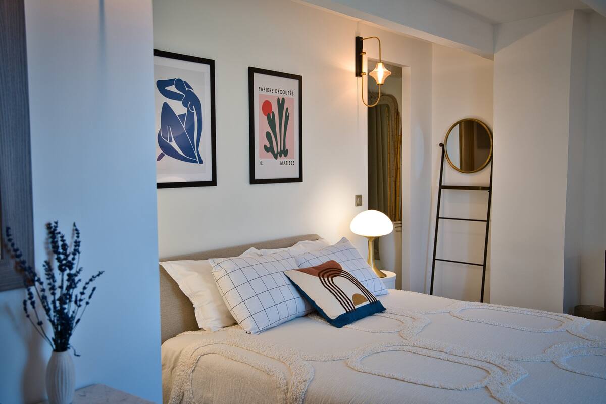 Quarto bem arrumado com cama, almofadas, mesa de cabeceira, um vaso com flores e obras de arte na parede.