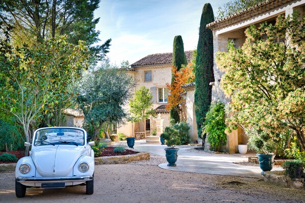 Une voiture ancienne est garée dans une allée, avec un bâtiment en pierre et une végétation luxuriante en arrière-plan.