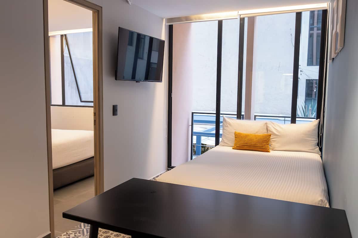 Una habitación de hotel moderna con cama, televisor de pantalla plana, balcón con barandilla y un ventanal grande.