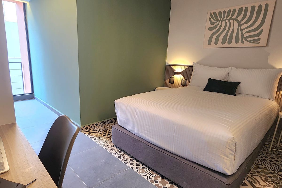 Habitación de hotel con una cama grande, un escritorio con silla, una mesita con planta y un adorno en la pared.