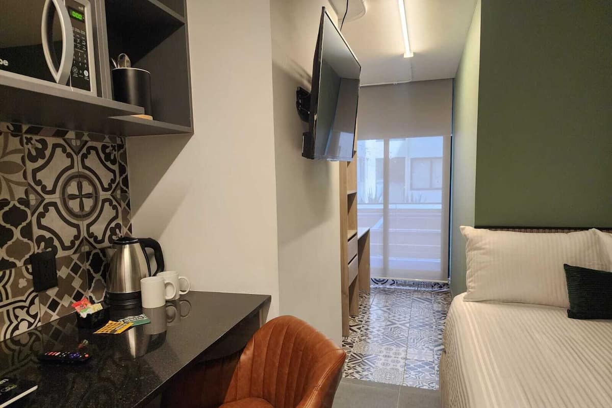 Una habitación de hotel con una cama, un escritorio con cafetera, una silla, un televisor montado en la pared y un suelo estampado.