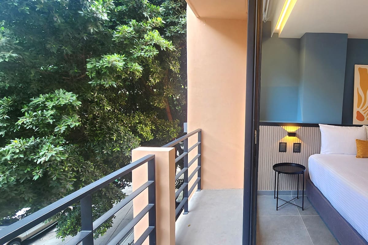 Un balcón con vistas a árboles, una cama con cabecera, una mesita y una lámpara fijada a la pared.