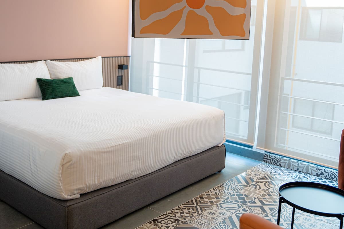 Habitación de hotel con una cama grande, un almohadón verde, una obra de arte moderna y abstracta, una pequeña mesa redonda y baldosas de suelo con diseño.