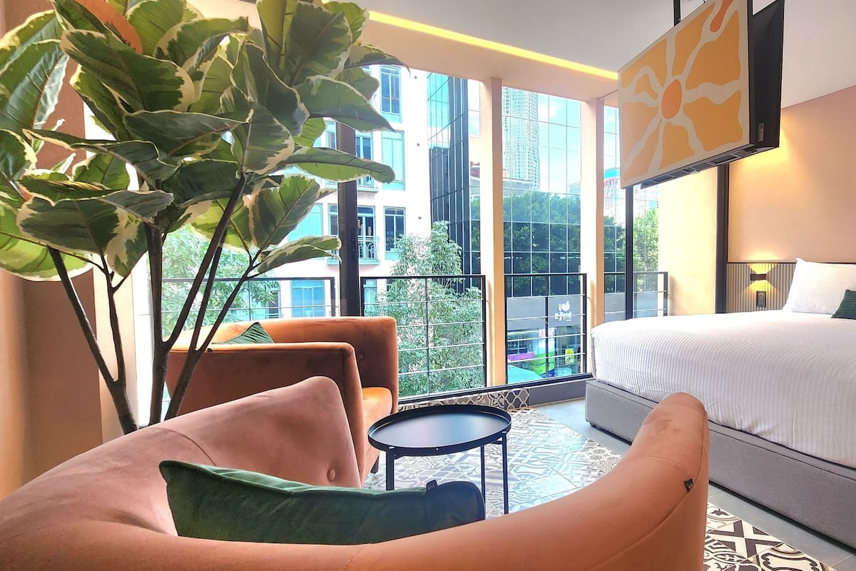 Una habitación de hotel moderna con una cama grande, una zona de estar con una silla y una mesita pequeña, y un ventanal que ofrece vistas de la ciudad.