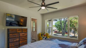 Une chambre à coucher comprenant un lit, un ventilateur de plafond, une télévision, une commode et une fenêtre donnant sur des arbres.