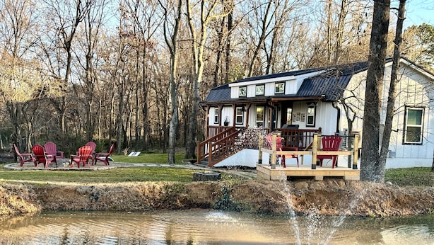 A lakeside cabin with a deck, red Adirondack chairs, and a small waterfall feature.