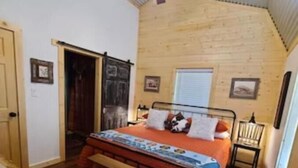 A bedroom with a wooden ceiling, a bed with a colorful blanket, a bench, and a fluffy rug.