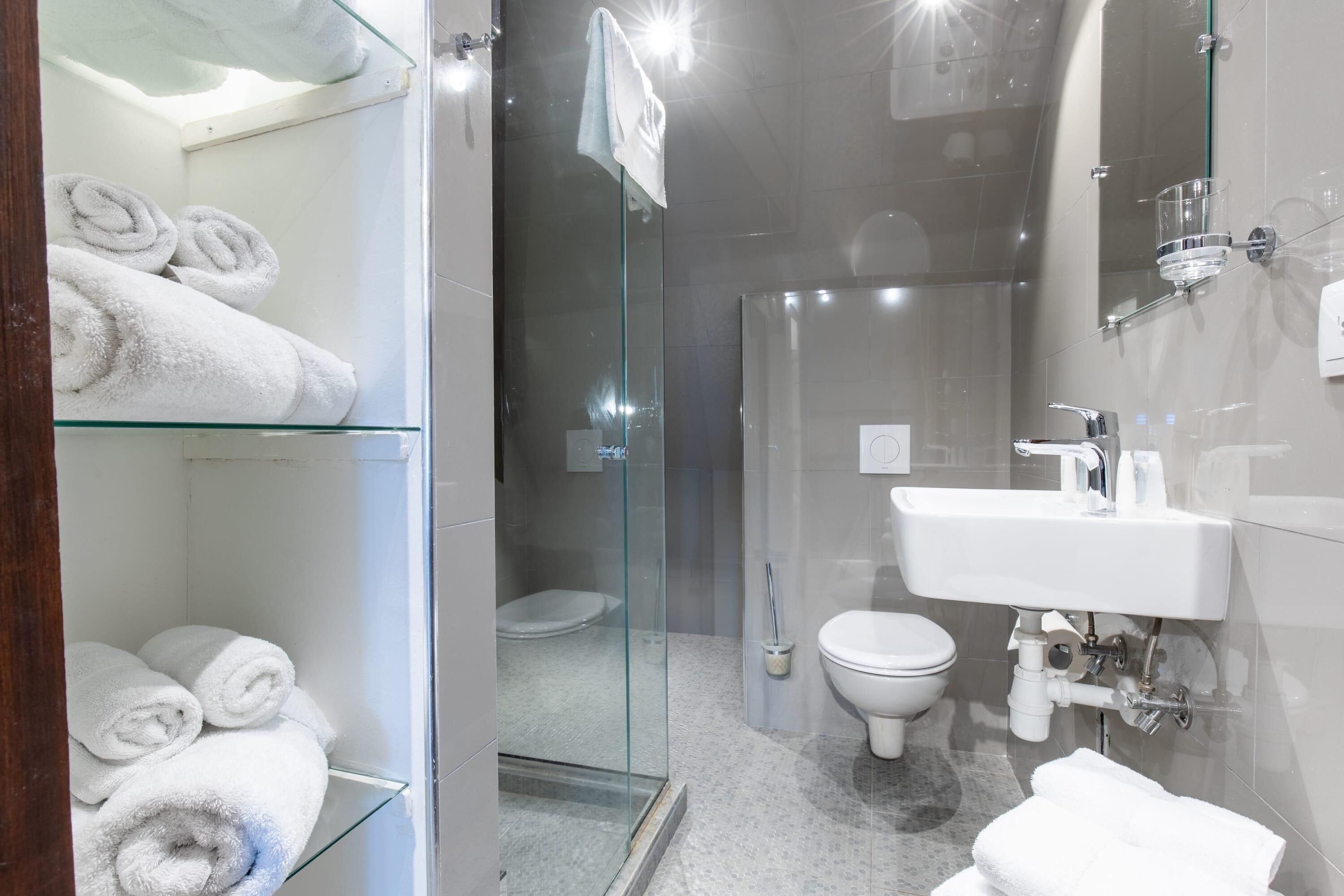 A modern bathroom with a glass shower enclosure, a white sink, and a glass shelf with rolled towels.