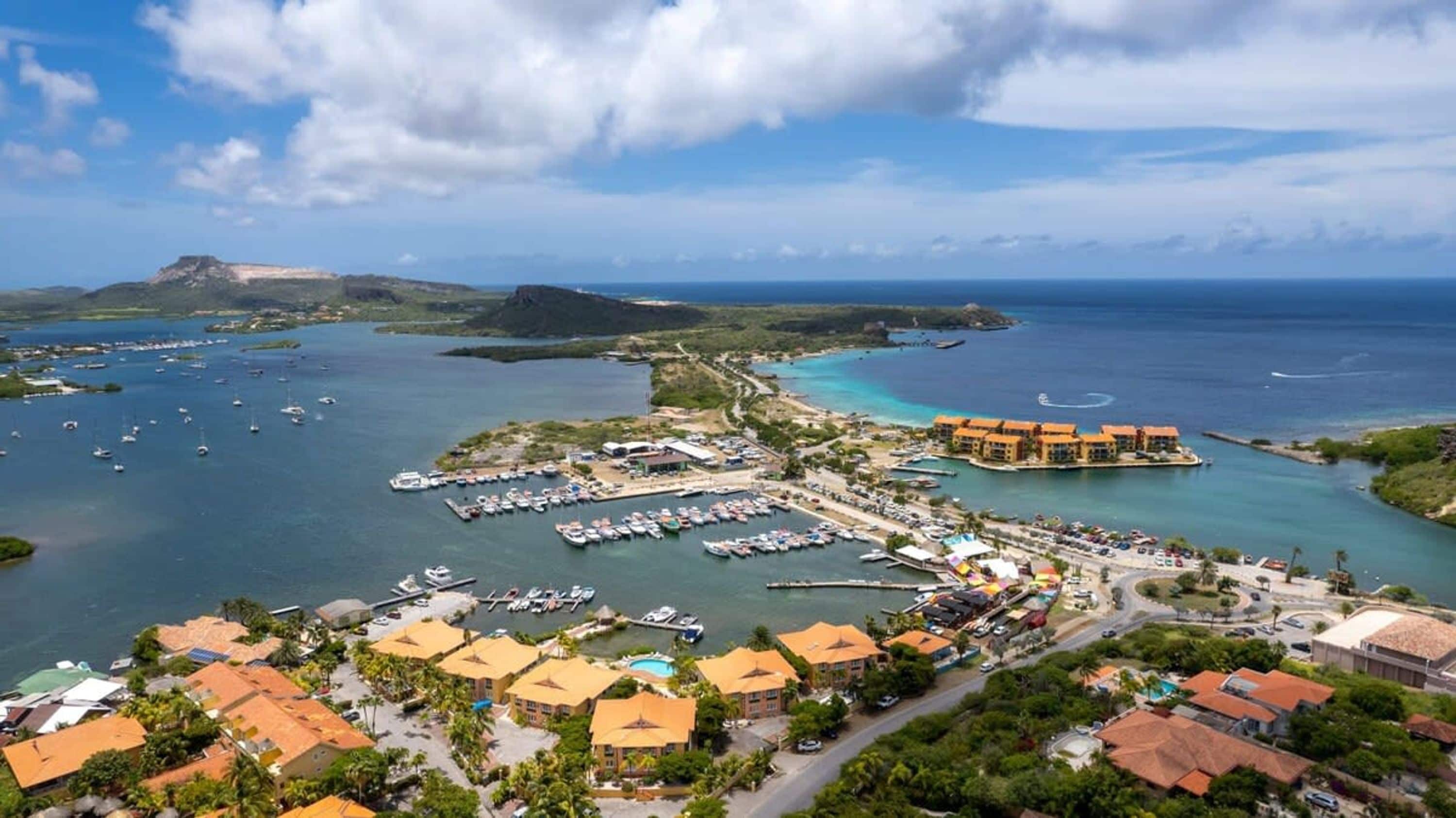 Vista aérea de un complejo turístico costero con varias edificaciones, un puerto deportivo con barcos y aguas azules cristalinas.