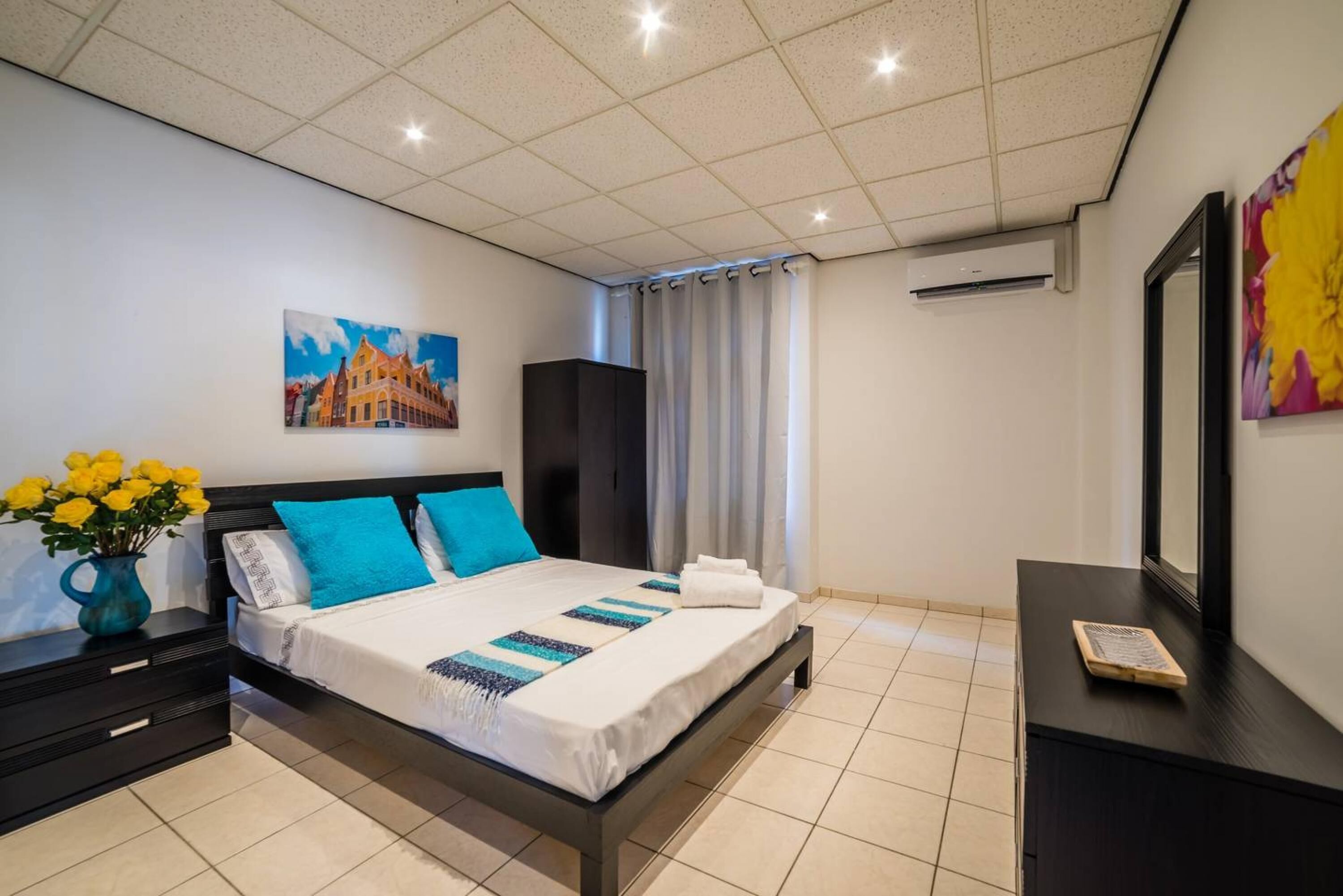 Un dormitorio moderno con una cama grande, una mesita de noche con un florero y un cuadro en la pared, además de un equipo de aire acondicionado.