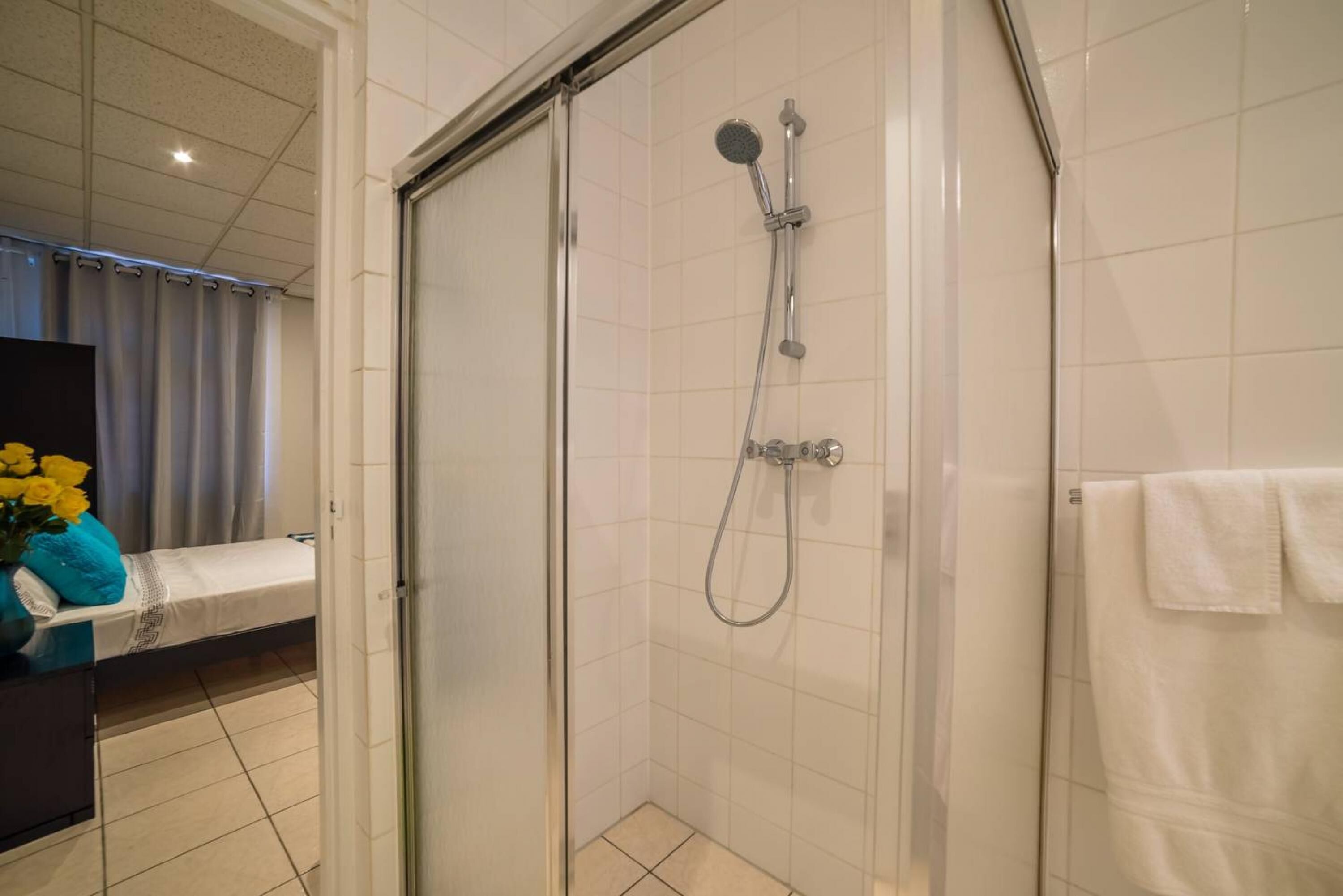Eine Dusche mit Handbrause und Wandarmatur, ein weißes Handtuch an der Wand und ein Bett mit blauem Kissen und einer Vase mit gelben Blumen.