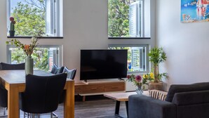 Ein modernes Wohnzimmer mit Flachbildfernseher, Holzmöbeln und großen Fenstern mit Blick auf Grünflächen und einem Segelboot-Gemälde.