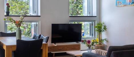 Un salón moderno con televisor de pantalla plana, muebles de madera y ventanas amplias que dan a vistas de vegetación y un cuadro de un velero.