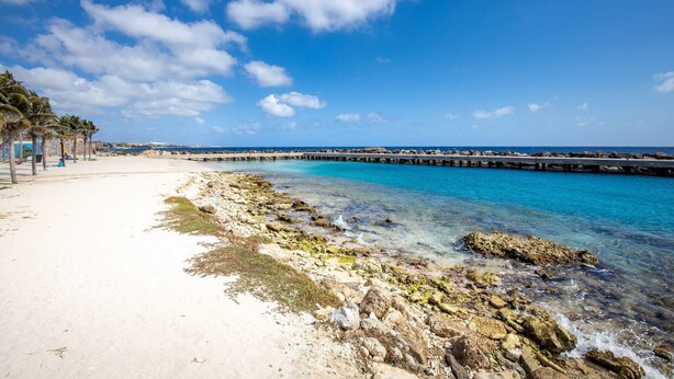 Ein Strand mit klarem, blauem Wasser, felsigem Ufer und einem Steg, der sich ins Meer erstreckt.