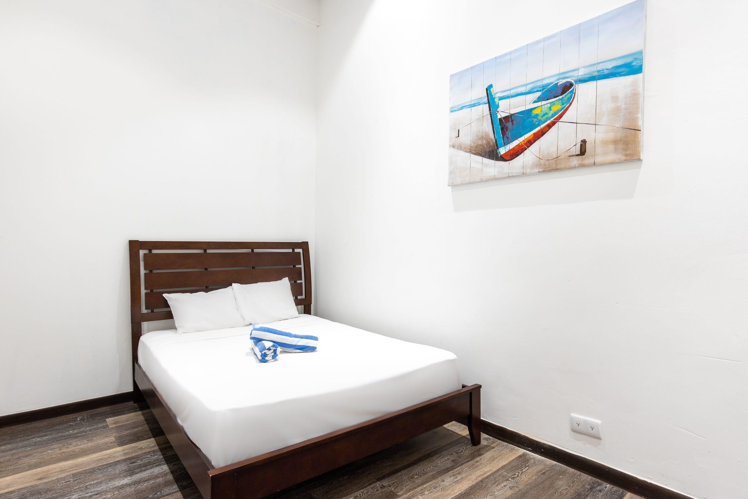 Una cama bien hecha con cabecera de madera, pared blanca y un cuadro de un barco en la pared.