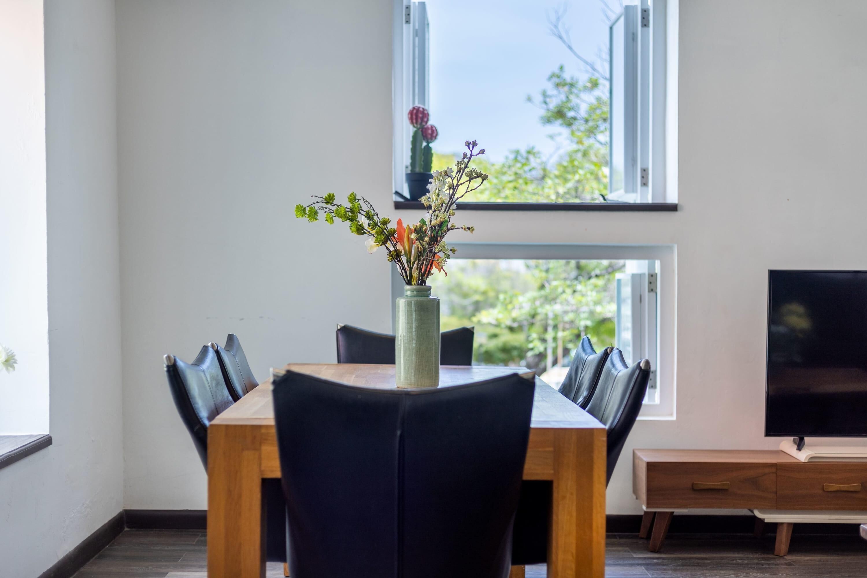 A dining area with a wooden table, black chairs, a vase with flowers, and a TV on a stand.