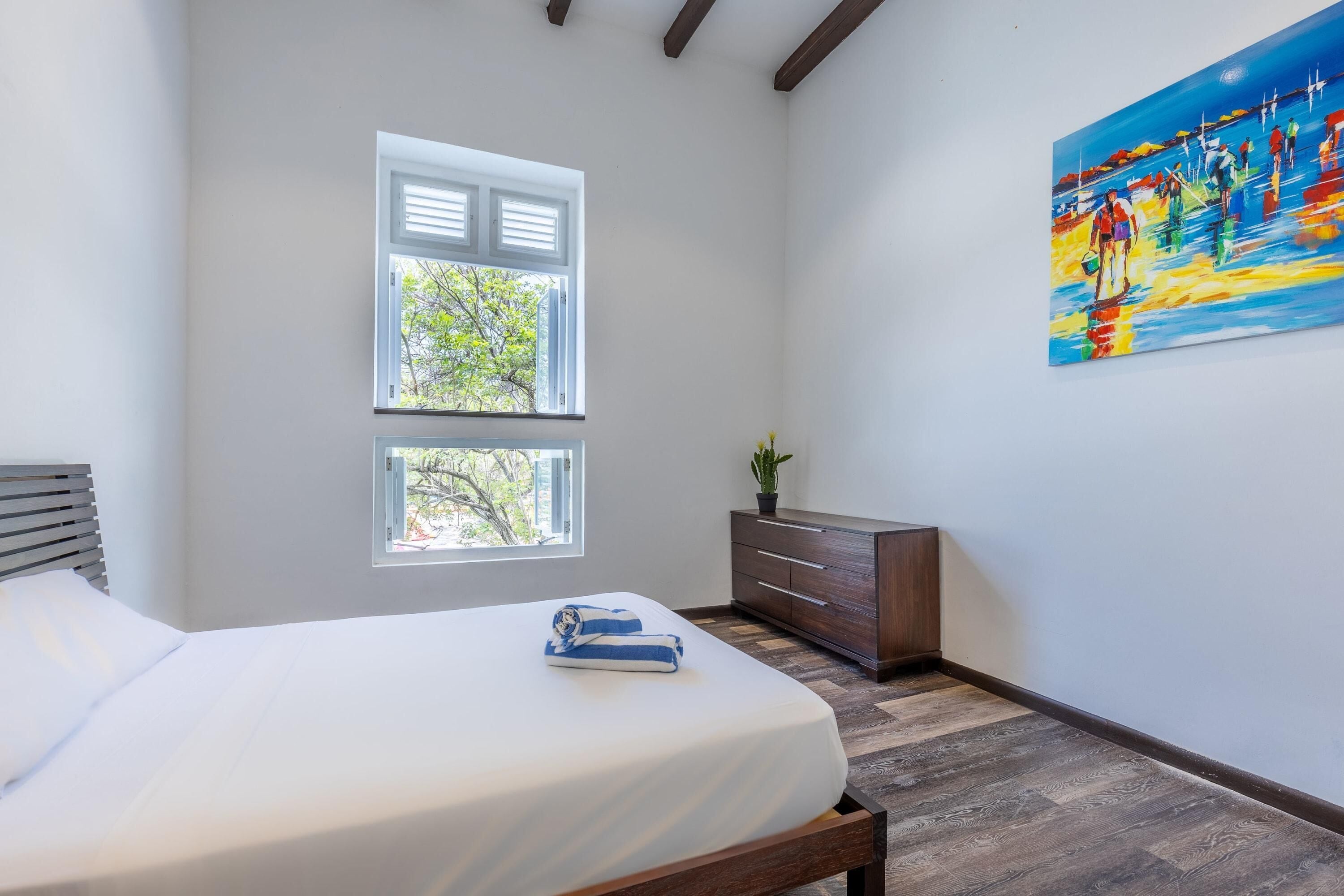A bedroom with a bed, two windows, a dresser, and a painting on the wall.