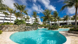 A swimming pool with a curved edge, surrounded by palm trees and a stone wall. - Blue Bay The Reef Balcony (Sint Michiel)