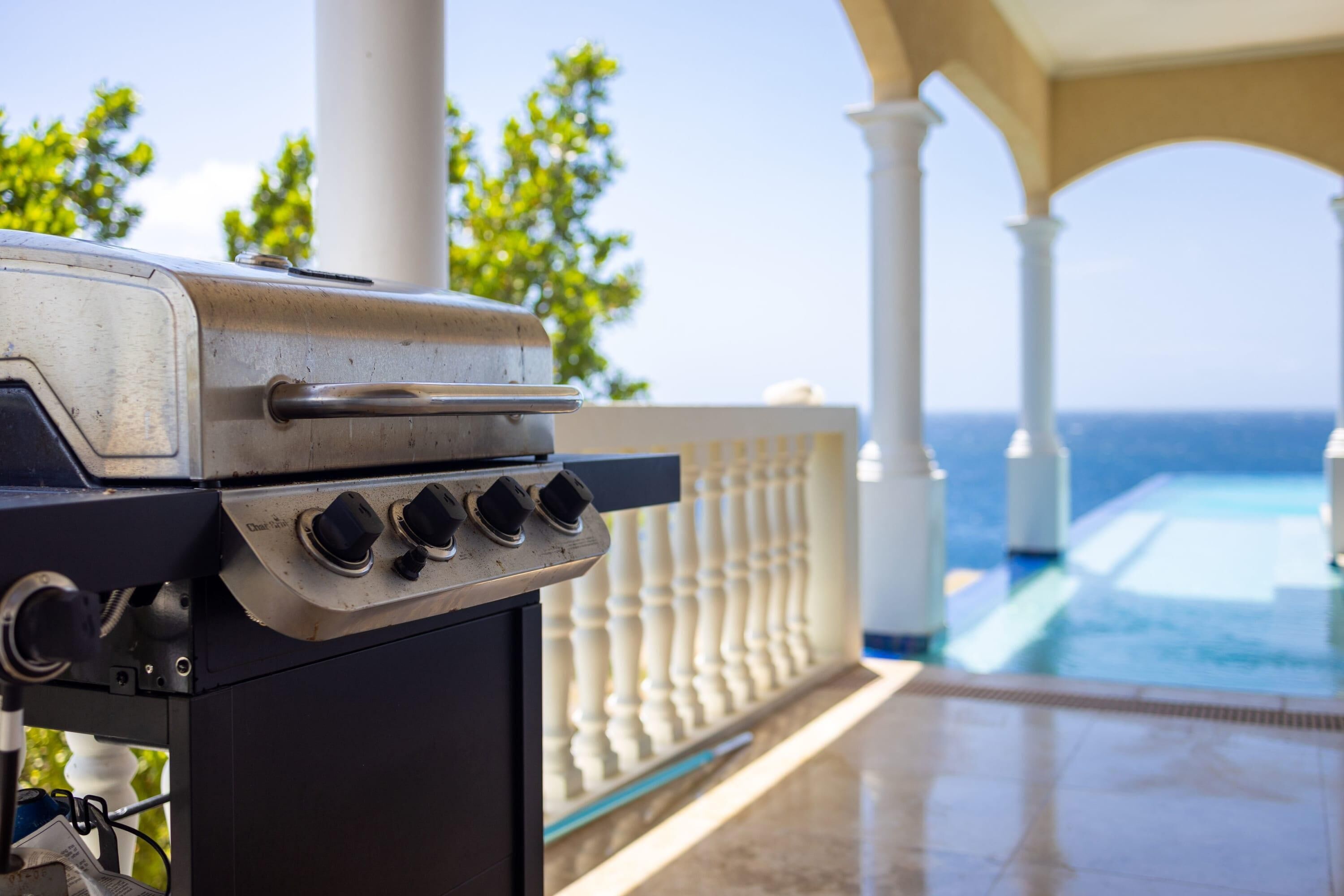 Una parrilla con cuatro quemadores y un asa, ubicada en un balcón con piscina y barandillas blancas al fondo.