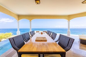 A long wooden table with wicker chairs, set against a view of the ocean. - Cliff Villa - Award Winning Luxury Retreat (Sabana Westpunt)