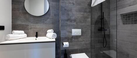 Une salle de bain moderne avec un grand miroir, un lavabo blanc, des toilettes et une douche séparée par une paroi de verre.