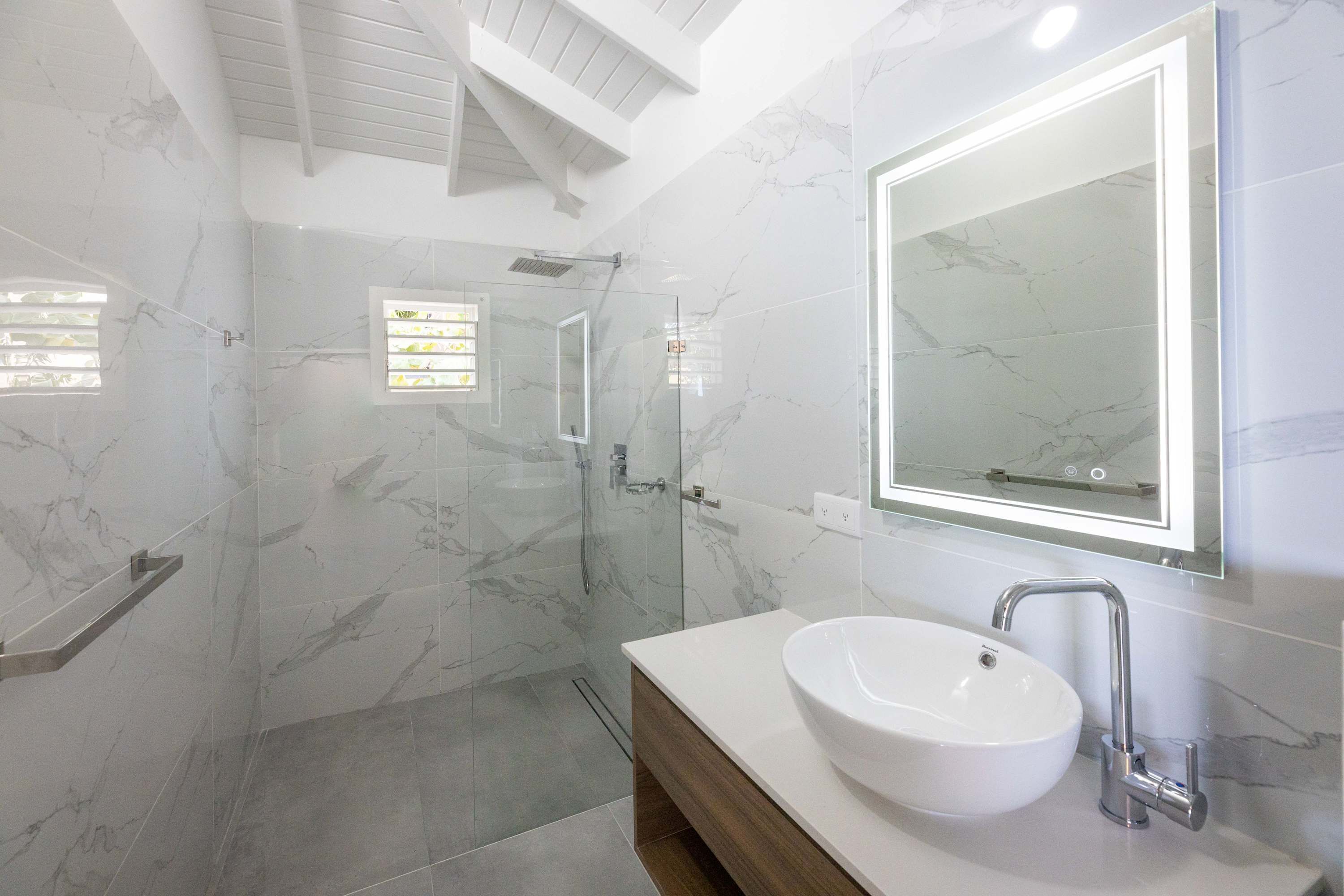 Un baño moderno con ducha de mármol, un espejo grande y un lavamanos blanco.