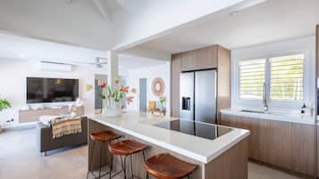 A modern kitchen with a central island, bar stools, a refrigerator, and a large window with blinds.