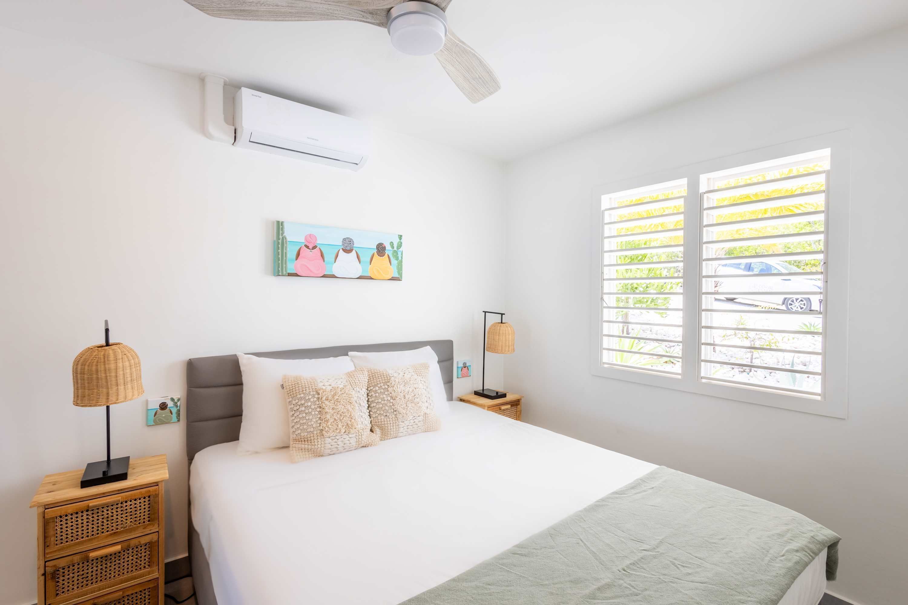 Ein Schlafzimmer mit einem Bett, zwei Nachttischen, einem Deckenventilator, einer Klimaanlage, einem Fenster mit Jalousien und einem Bild an der Wand.