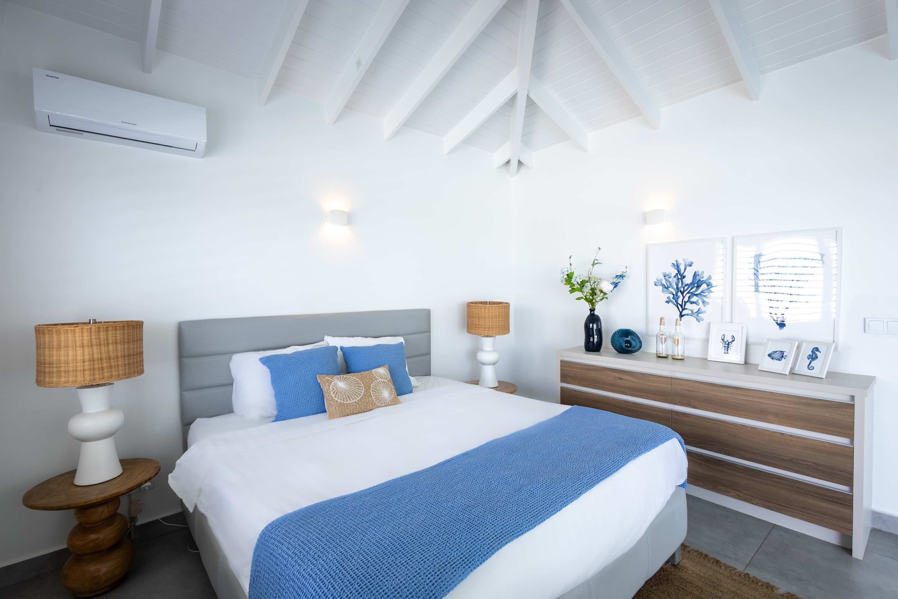 A bedroom with a bed, bedside lamps, a bedside table, a nightstand, a dresser, a window with blinds, and an air conditioning unit.