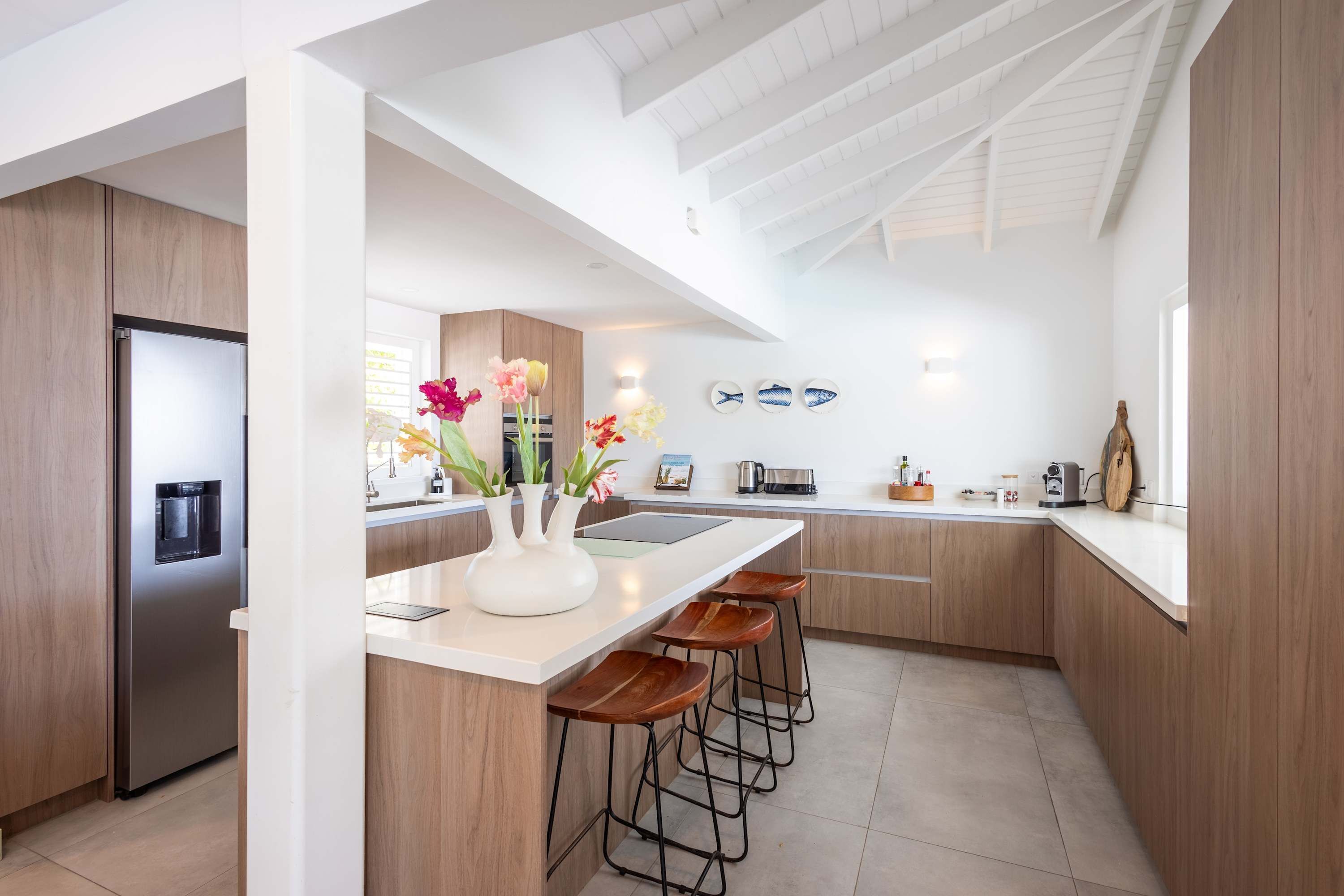 A modern kitchen with a central island, bar stools, a stainless steel refrigerator, and decorative fish wall art.