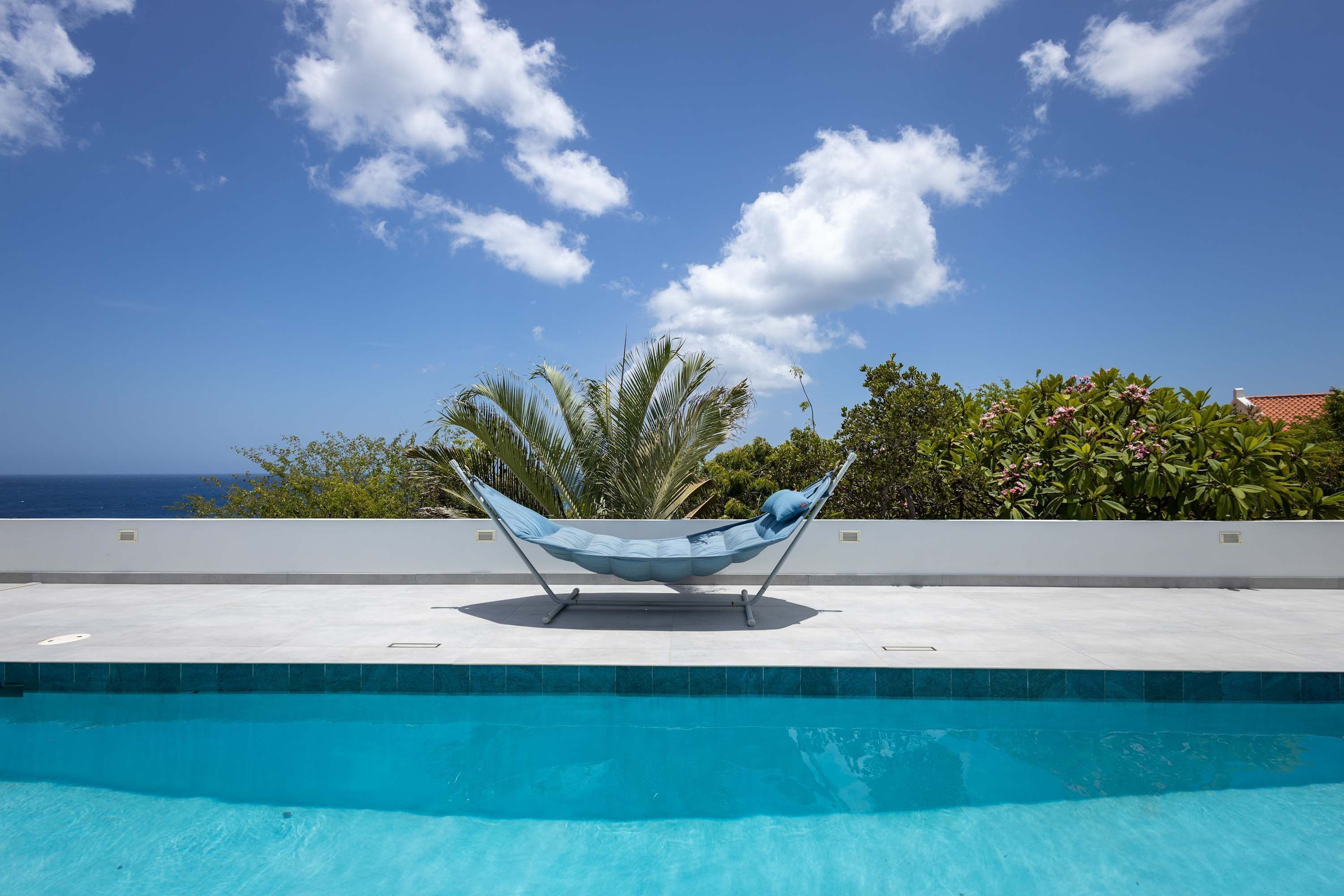Un'amaca vicino a una piscina con vista sull'oceano e un muro bianco con vegetazione.