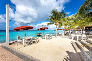 Een restaurant aan het strand met terras, palmbomen en uitzicht op zee.