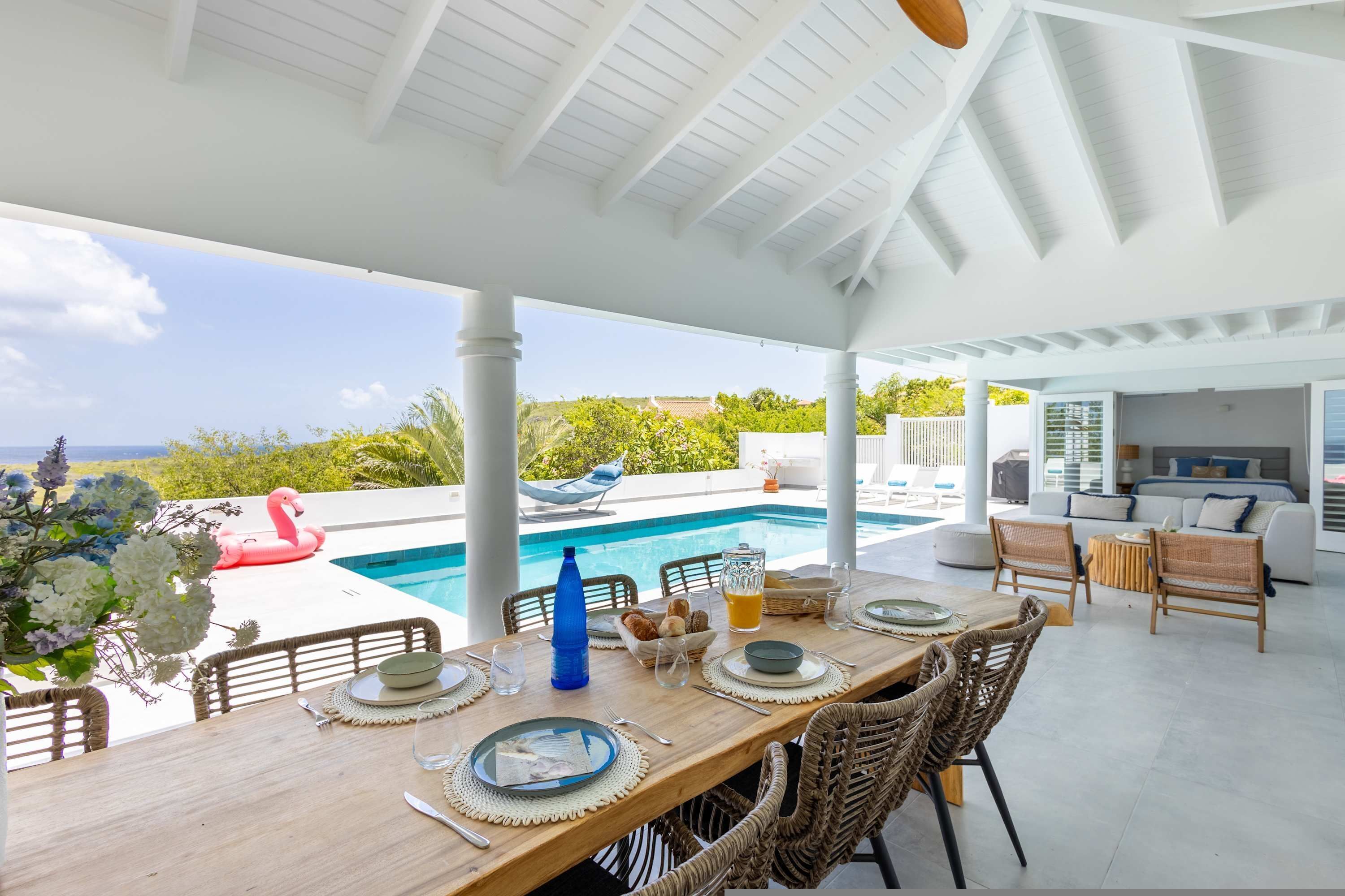 A covered outdoor dining area with a pool, wicker furniture, and a view of the ocean.