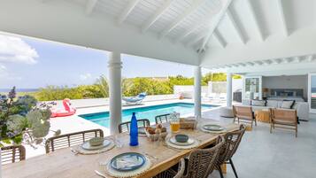 A covered outdoor dining area with a pool, wicker furniture, and a view of the ocean.