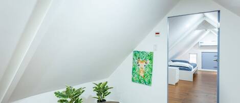 A room with a sloped ceiling, wooden flooring, a round table with two potted plants, and a painting of a giraffe on the wall.