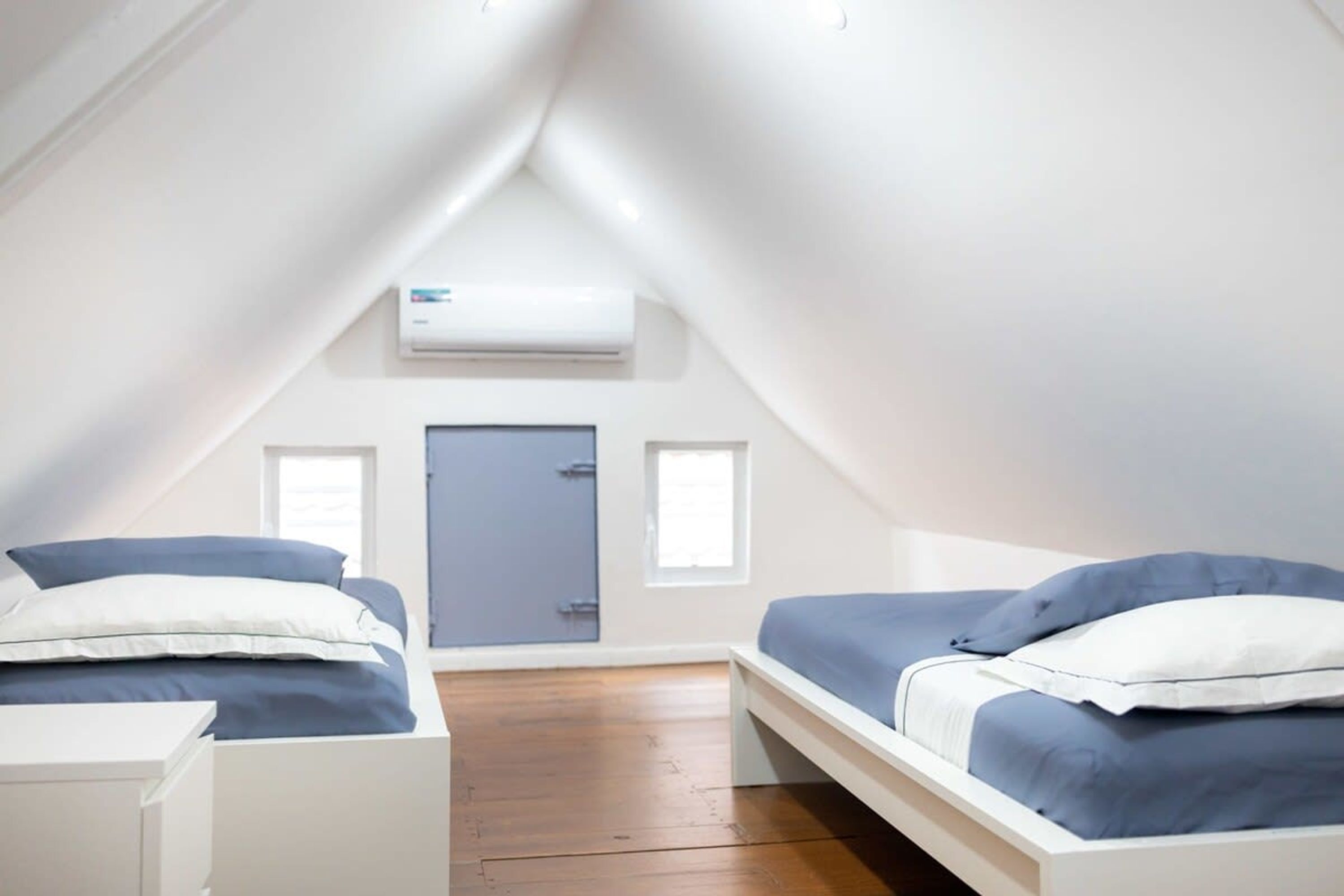 A small, modern room with two beds, a white desk, and an air conditioning unit on the wall.