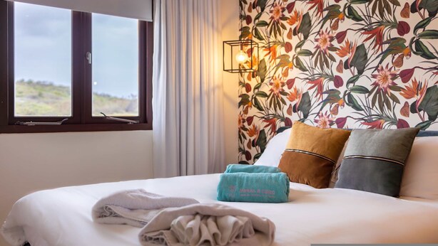Une chambre d’hôtel avec un lit, une fenêtre avec des rideaux, un papier peint à motifs floraux et un luminaire fixé au mur.