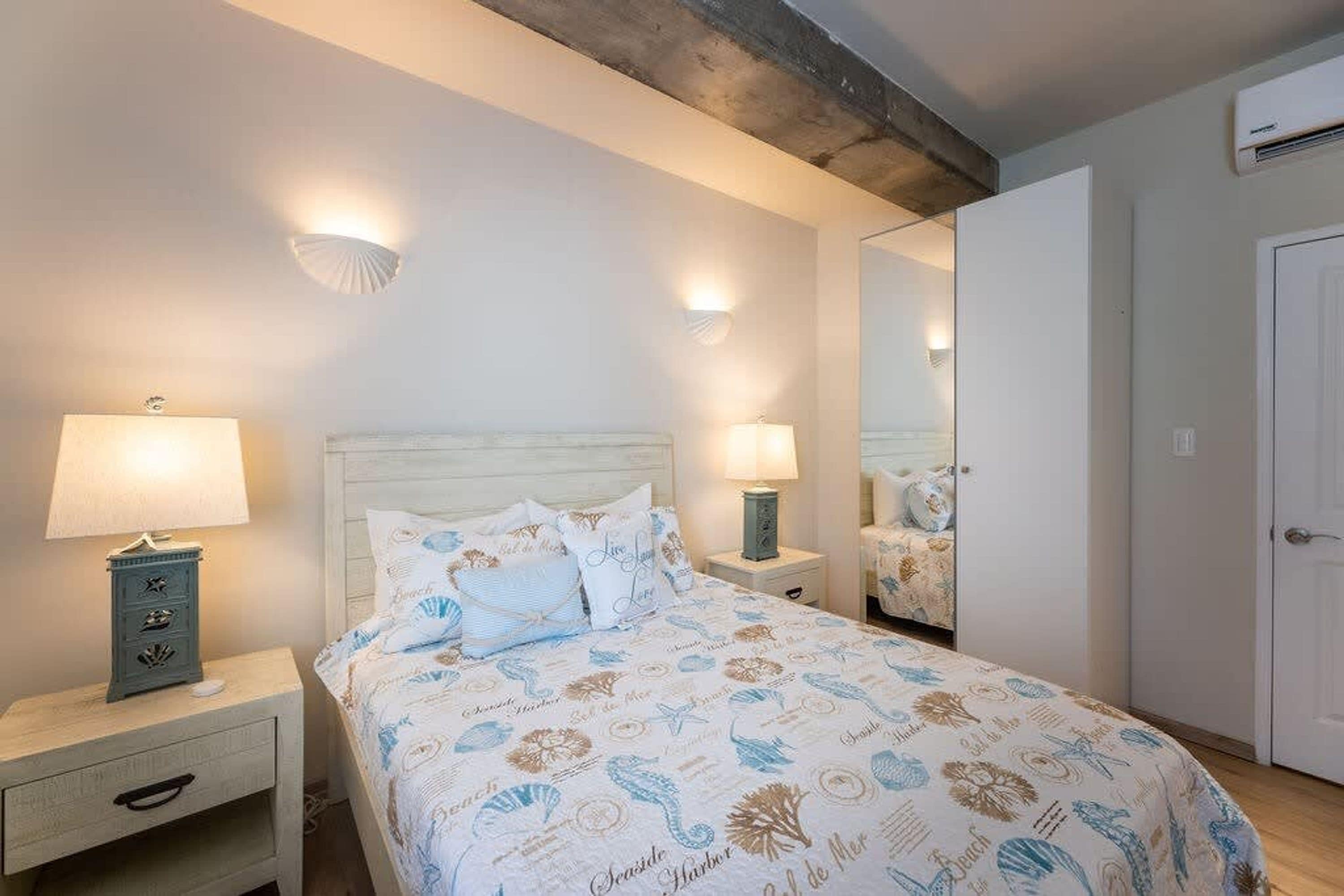 A bedroom with a bed, two bedside lamps, a nightstand with a drawer, a mirror, and a door.