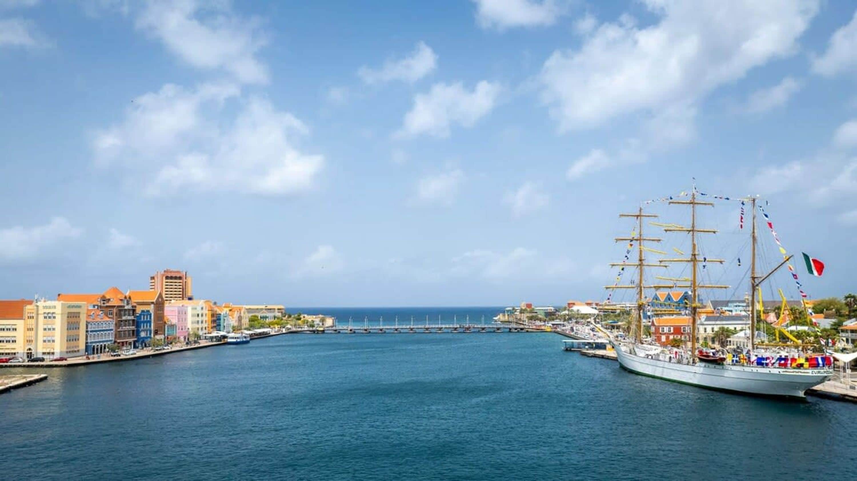 A harbor with a tall ship docked, a clear sky, and buildings along the waterfront.