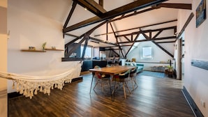 Un loft espacioso con vigas de madera a la vista, una hamaca, una mesa de comedor de madera y una cocina al fondo.