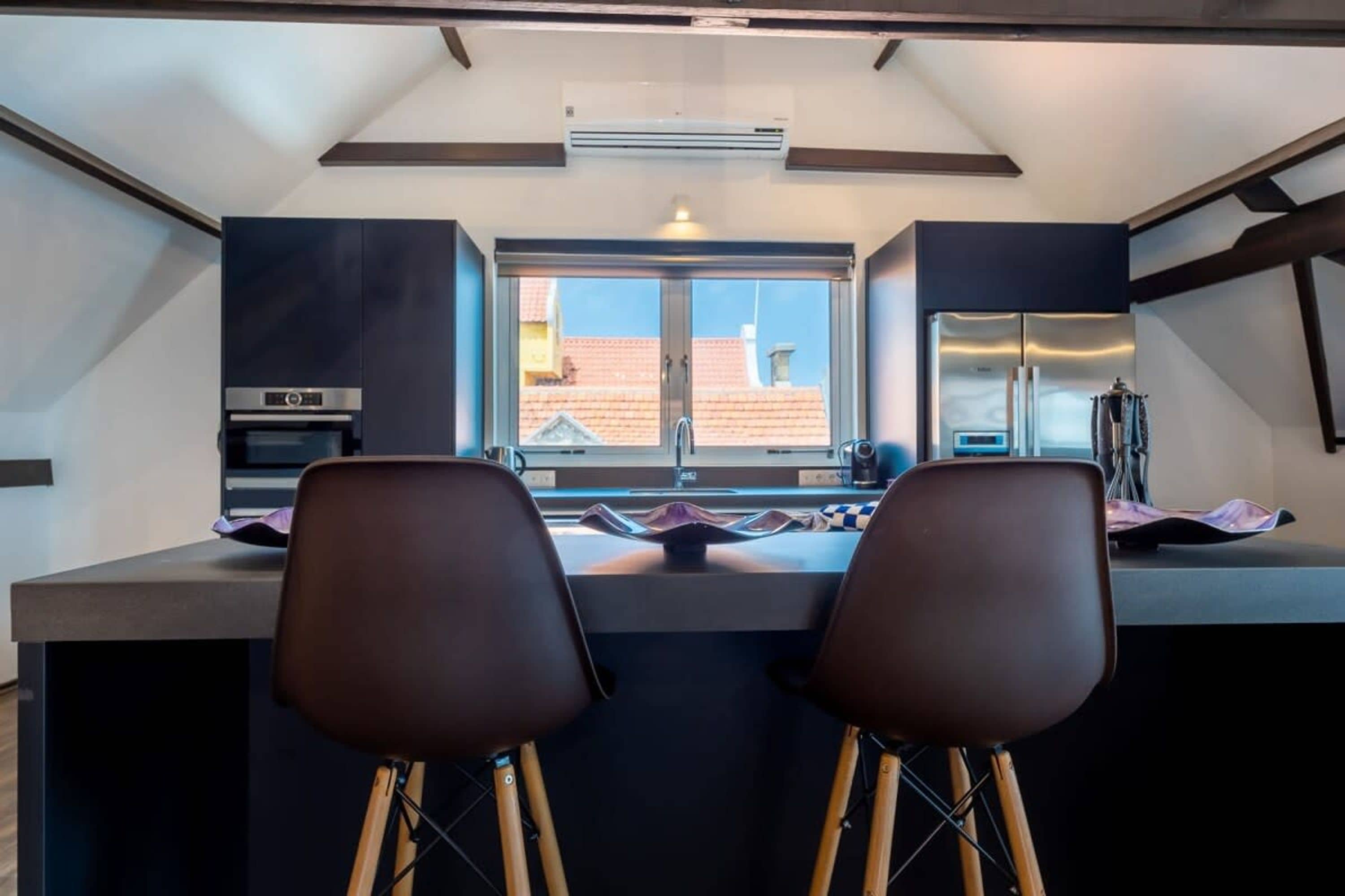 A modern kitchen with a central island, two bar stools, and a large window providing a view of the cityscape.