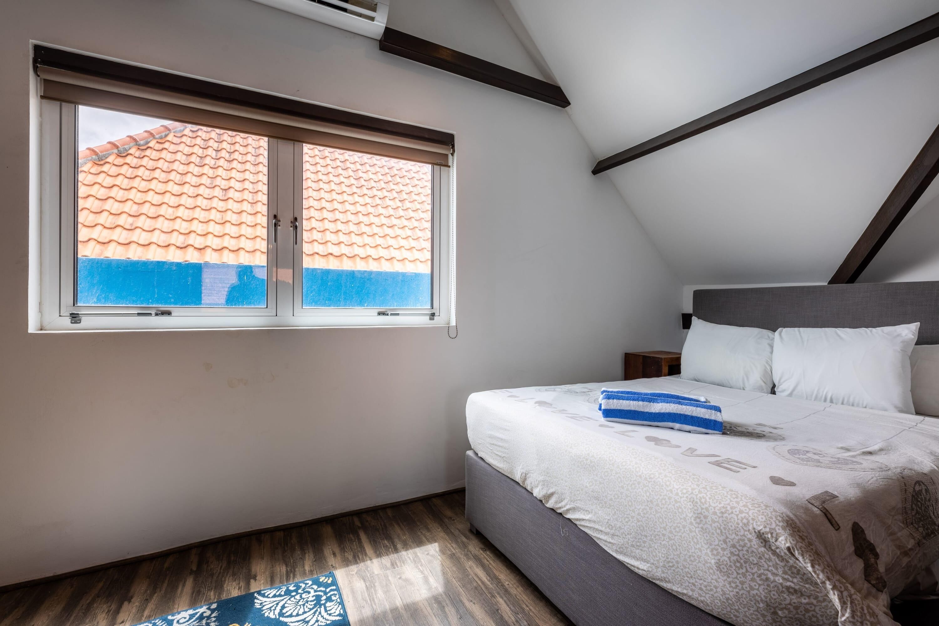 A bedroom with a sloped ceiling, a large window with a view of a tiled roof, a bed with white bedding, and a small bedside table.
