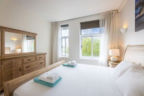 A hotel room with a large bed, a wooden dresser, a mirror, two windows with curtains, and a nightstand with a lamp. - Heart of the City Plaza Jojo Heritage Home (Willemstad)
