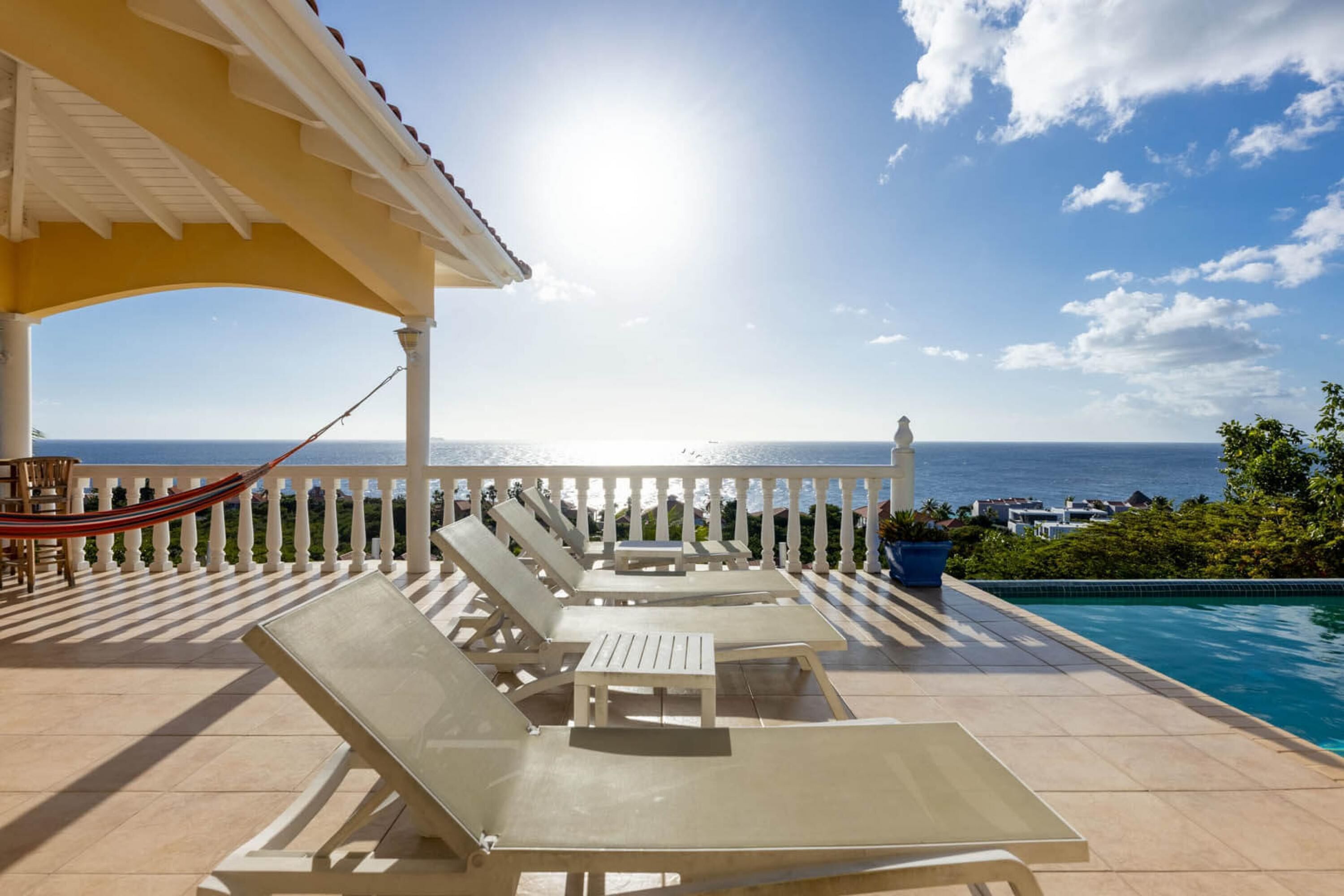 A sunlit outdoor lounge area with white lounge chairs and a pool, overlooking a clear blue sea.