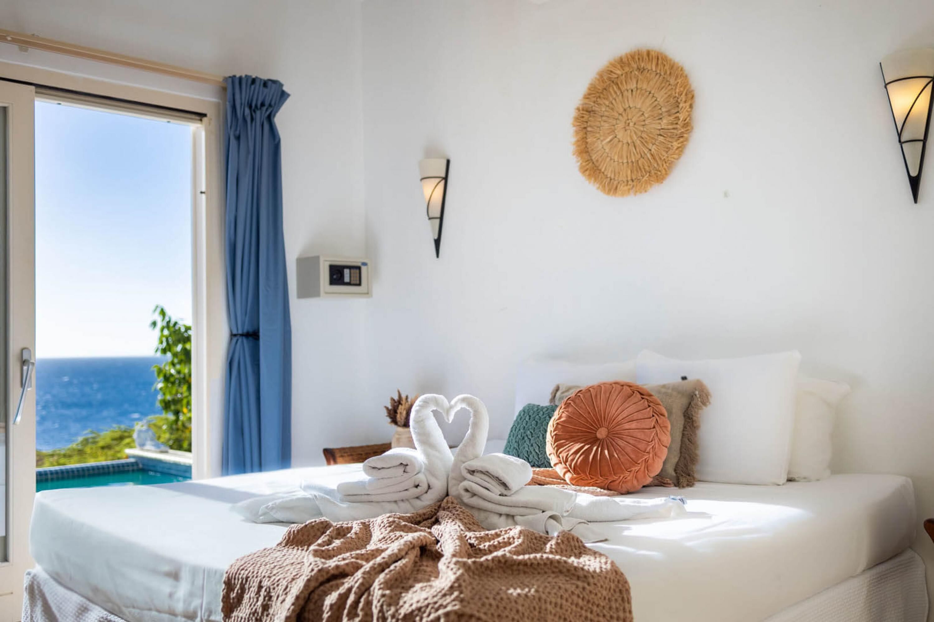 A bedroom with a bed, a view of the sea, a woven wall decoration, and a wall-mounted light fixture.