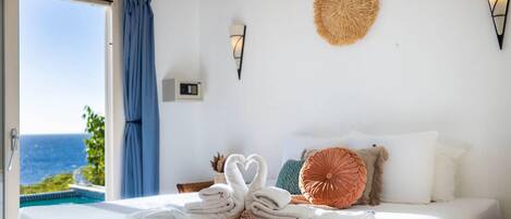 A bedroom with a bed, a view of the sea, a woven wall decoration, and a wall-mounted light fixture.